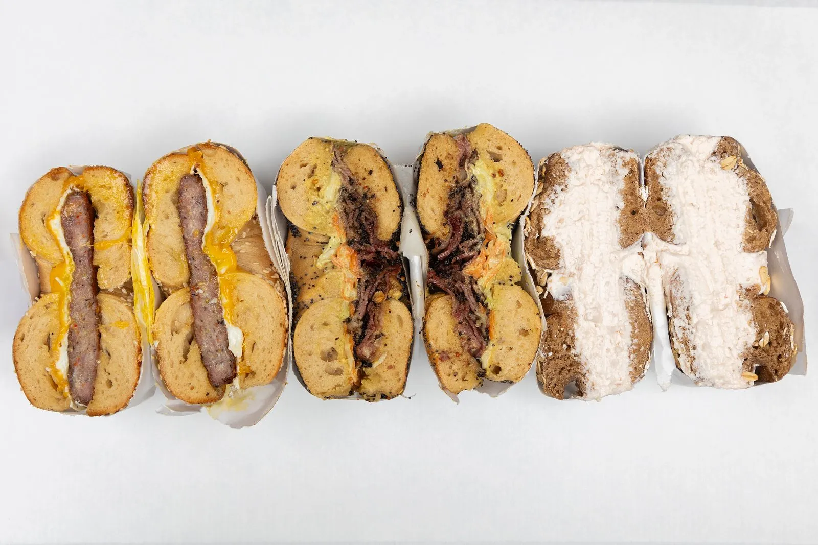 A row of five Gotham bagel sandwiches cut in half on a white table, from sausage and egg to cream cheese