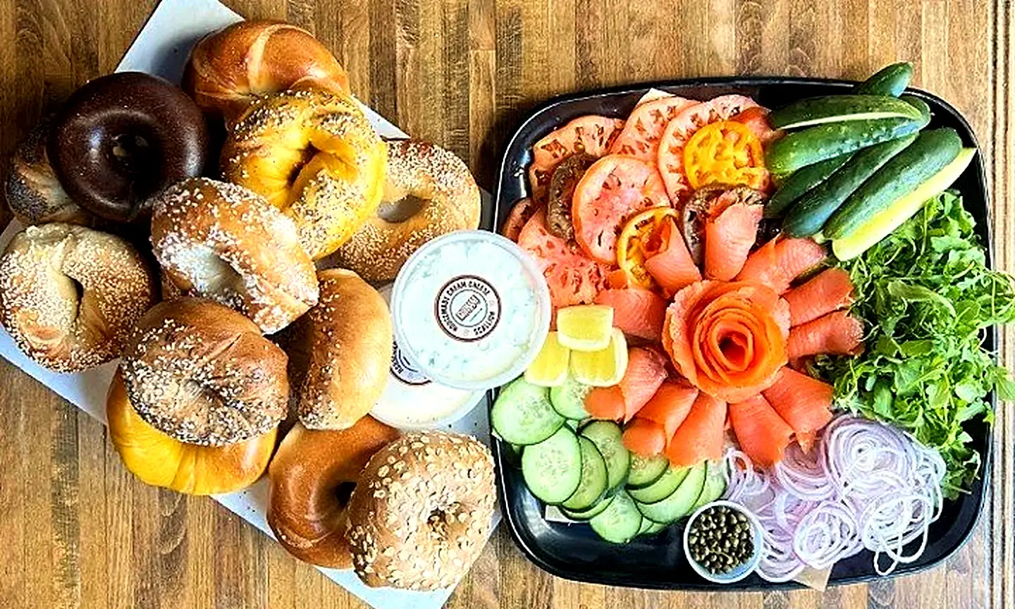 A dozen assorted hand-rolled bagels beside a lox platter with tomato, cucumber, arugula, red onion and capers