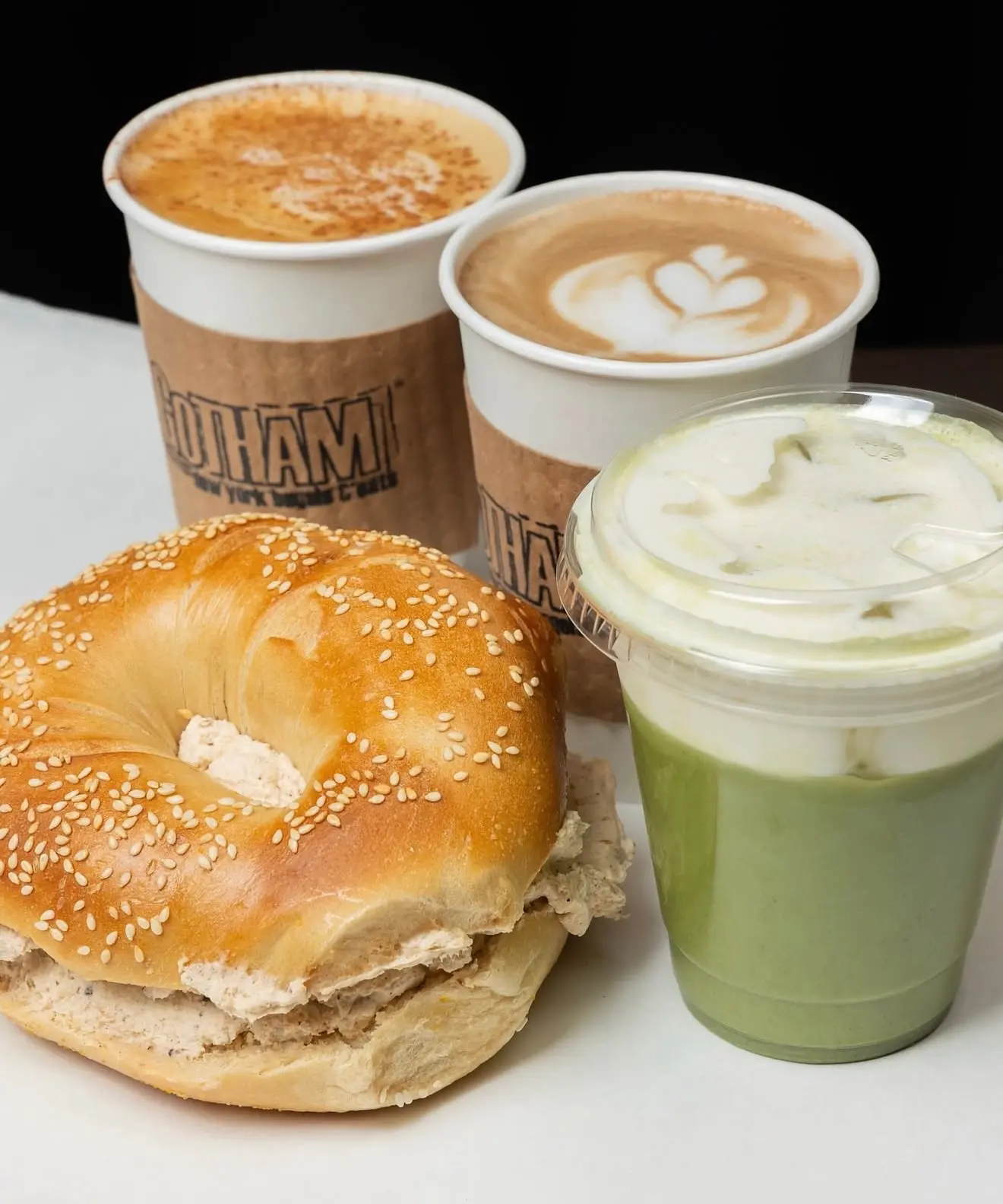 An iced Sweet Cream Matcha Latte with a thick cream top, beside two hot lattes in Gotham sleeves and a sesame bagel with cream cheese