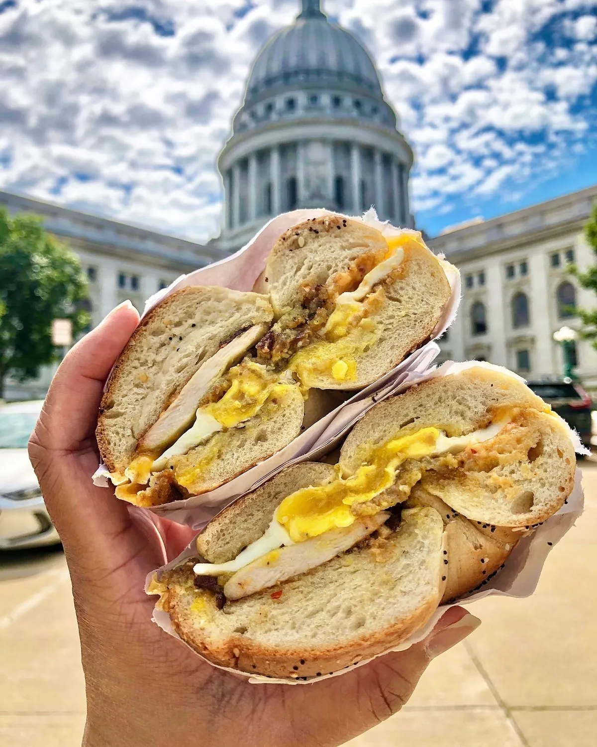 A Gotham Madison egg sandwich on a hand-rolled bagel, held up in front of the Wisconsin State Capitol