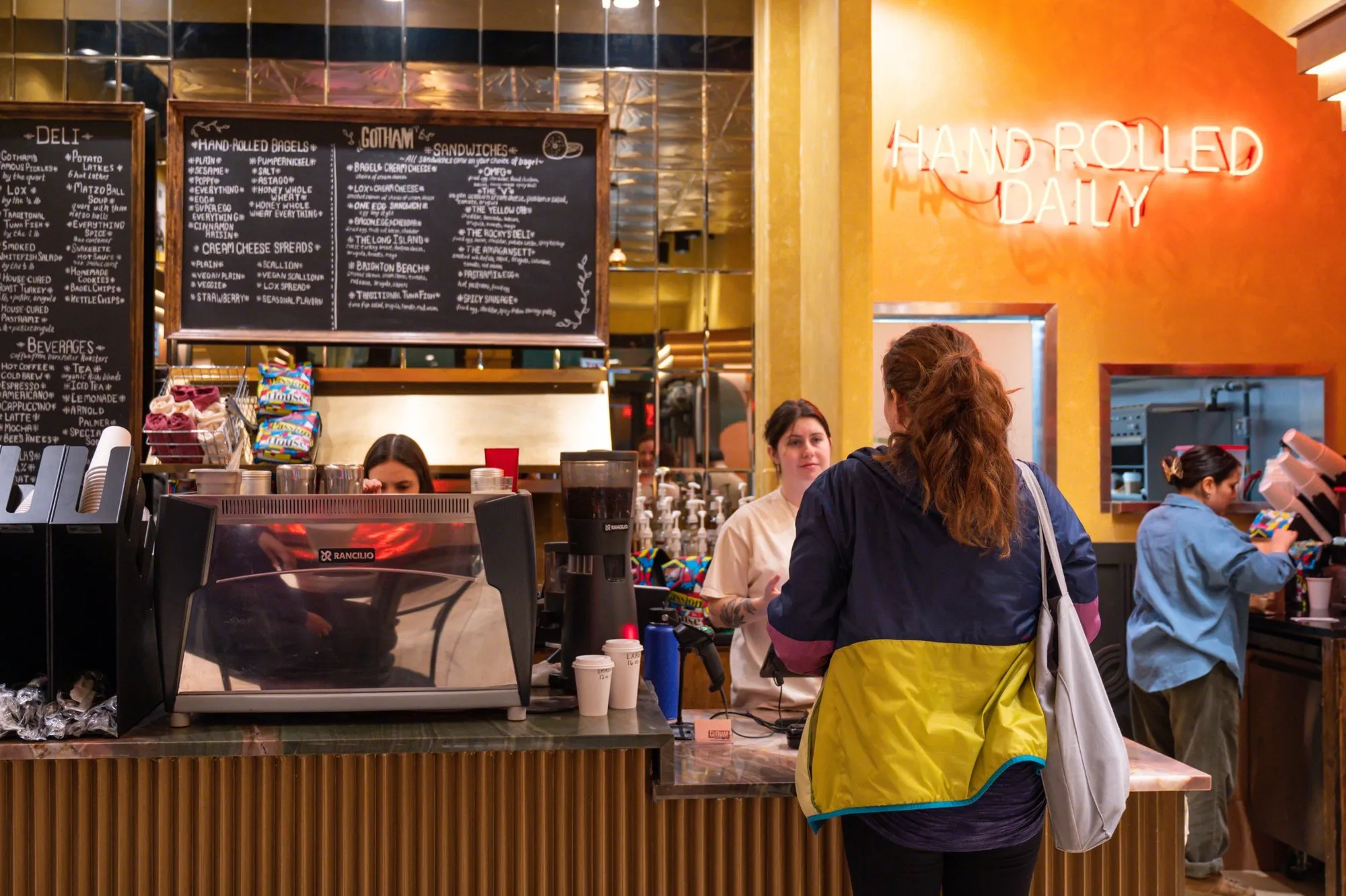 The counter at Gotham Bagels Magnificent Mile: chalkboard menus, an espresso machine and Hand Rolled Daily neon on an orange wall