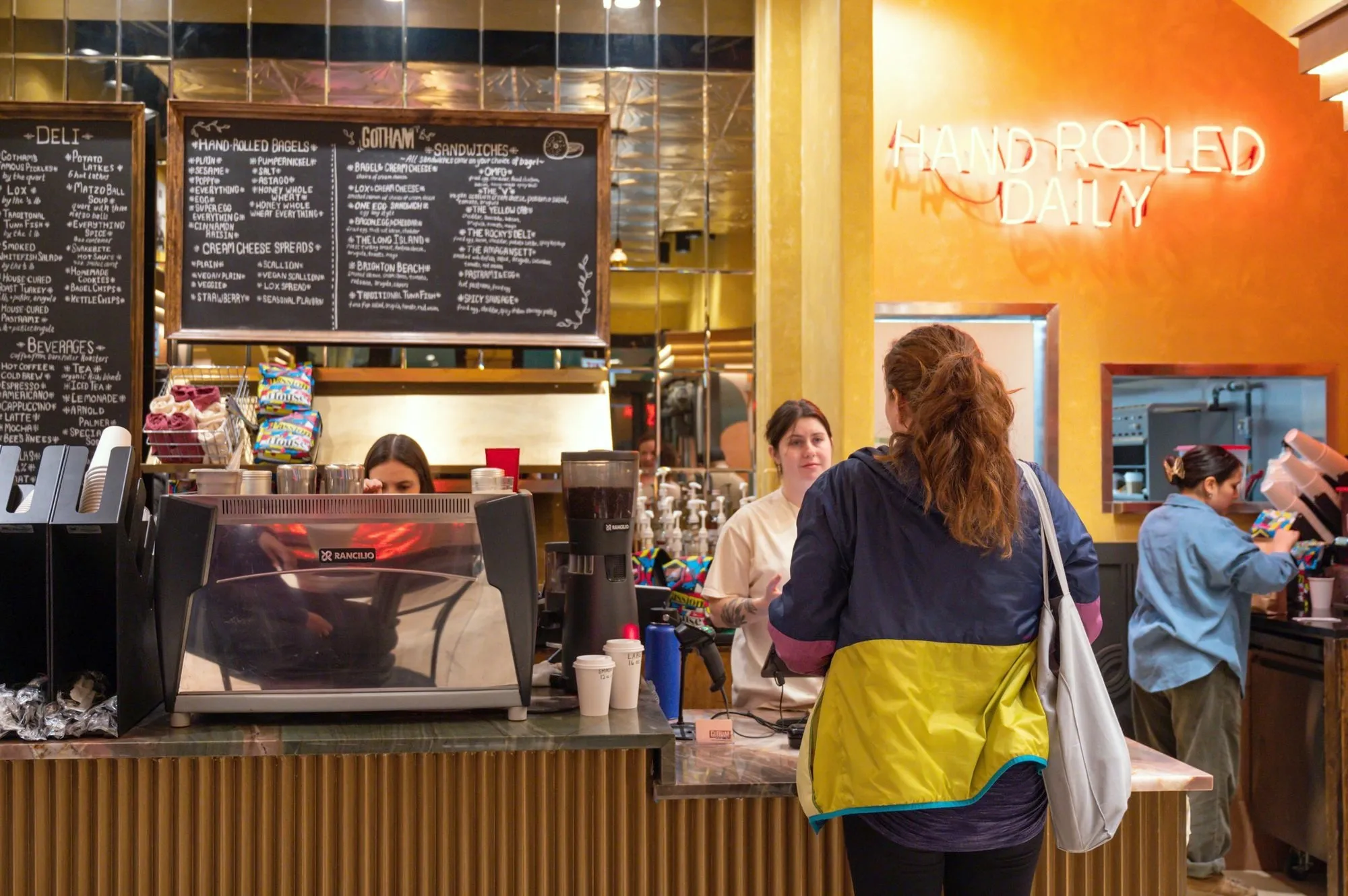 The counter at Gotham Bagels Magnificent Mile: chalkboard menus, an espresso machine and Hand Rolled Daily neon on an orange wall