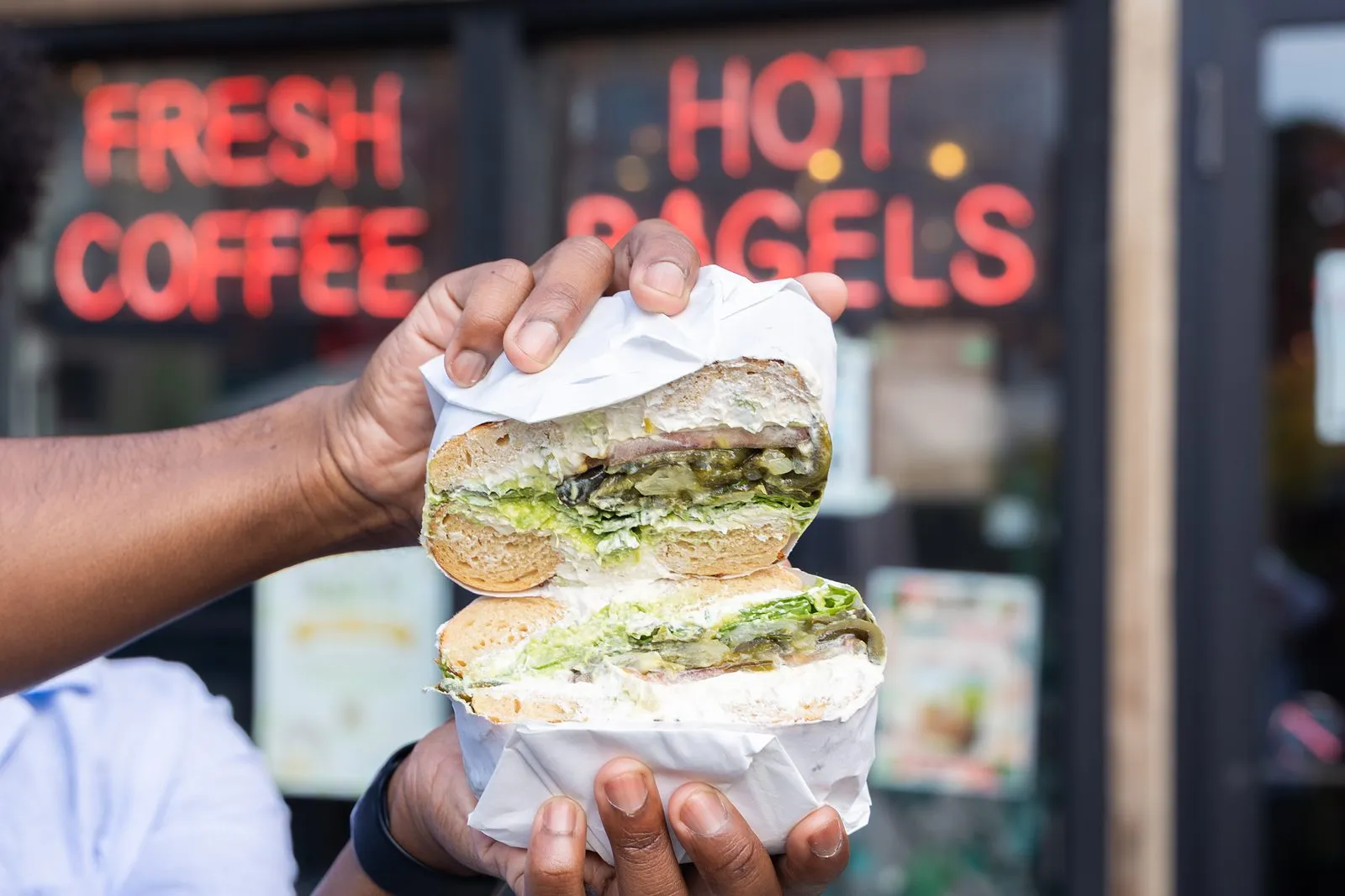 A Gotham bagel sandwich held up in front of the Fresh Coffee and Hot Bagels neon sign