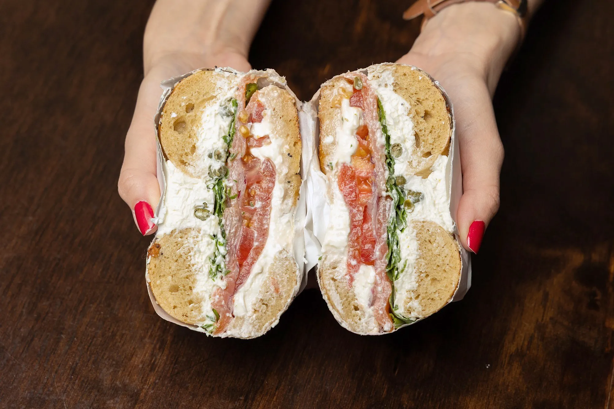 A Brighton Beach sandwich with lox, cream cheese, tomato and capers, cut in half and held in two hands