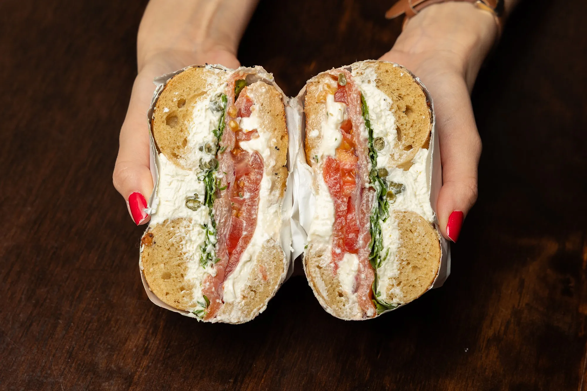 A Brighton Beach sandwich with lox, cream cheese, tomato and capers, cut in half and held in two hands