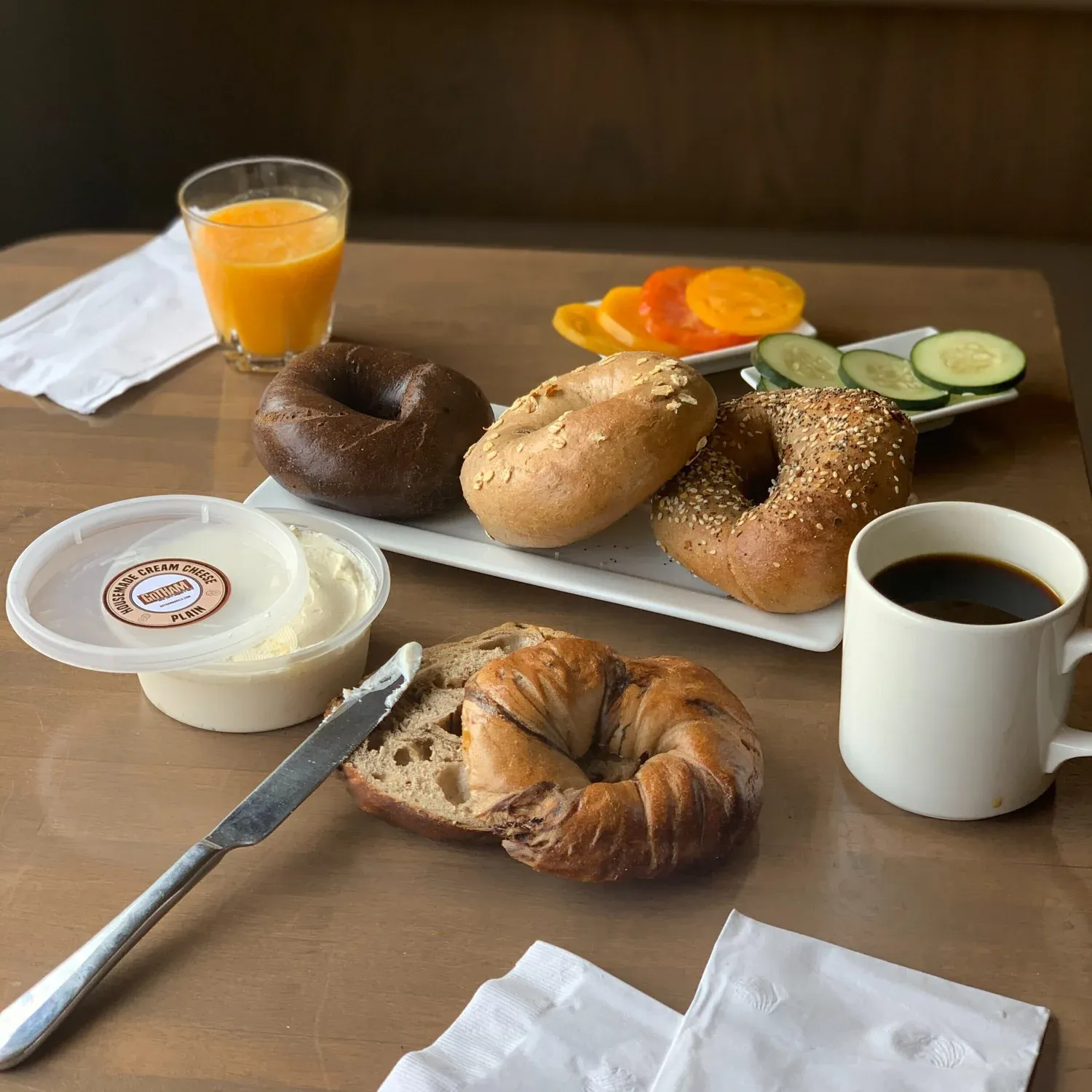 Breakfast spread of bagels, a tub of plain cream cheese, orange juice, black coffee and sliced tomato and cucumber