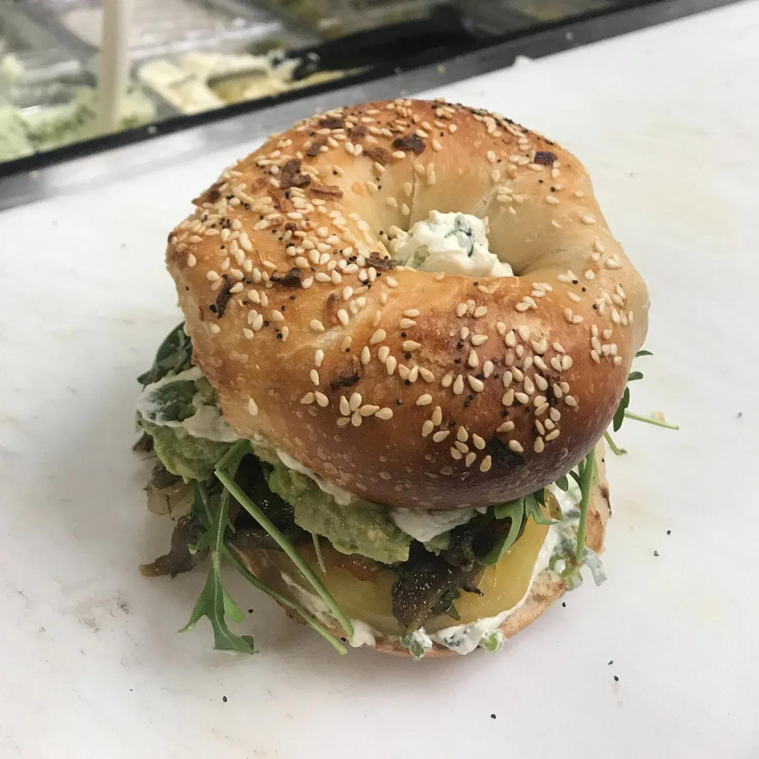 Everything bagel sandwich stacked with guacamole, caramelized onions, tomato, arugula and a herbed spread on a marble counter