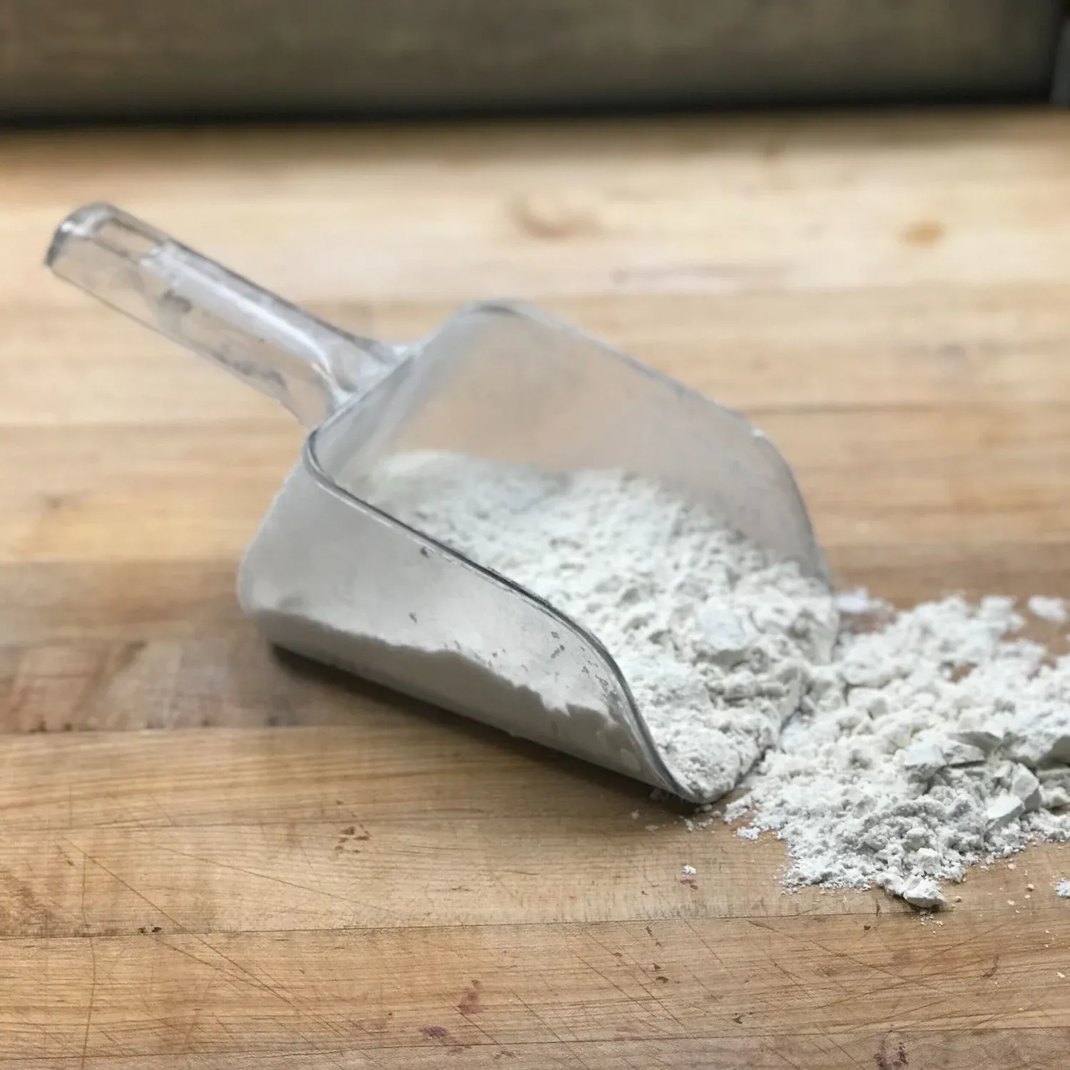 Clear plastic scoop spilling a pile of white flour onto a worn wooden bakery bench