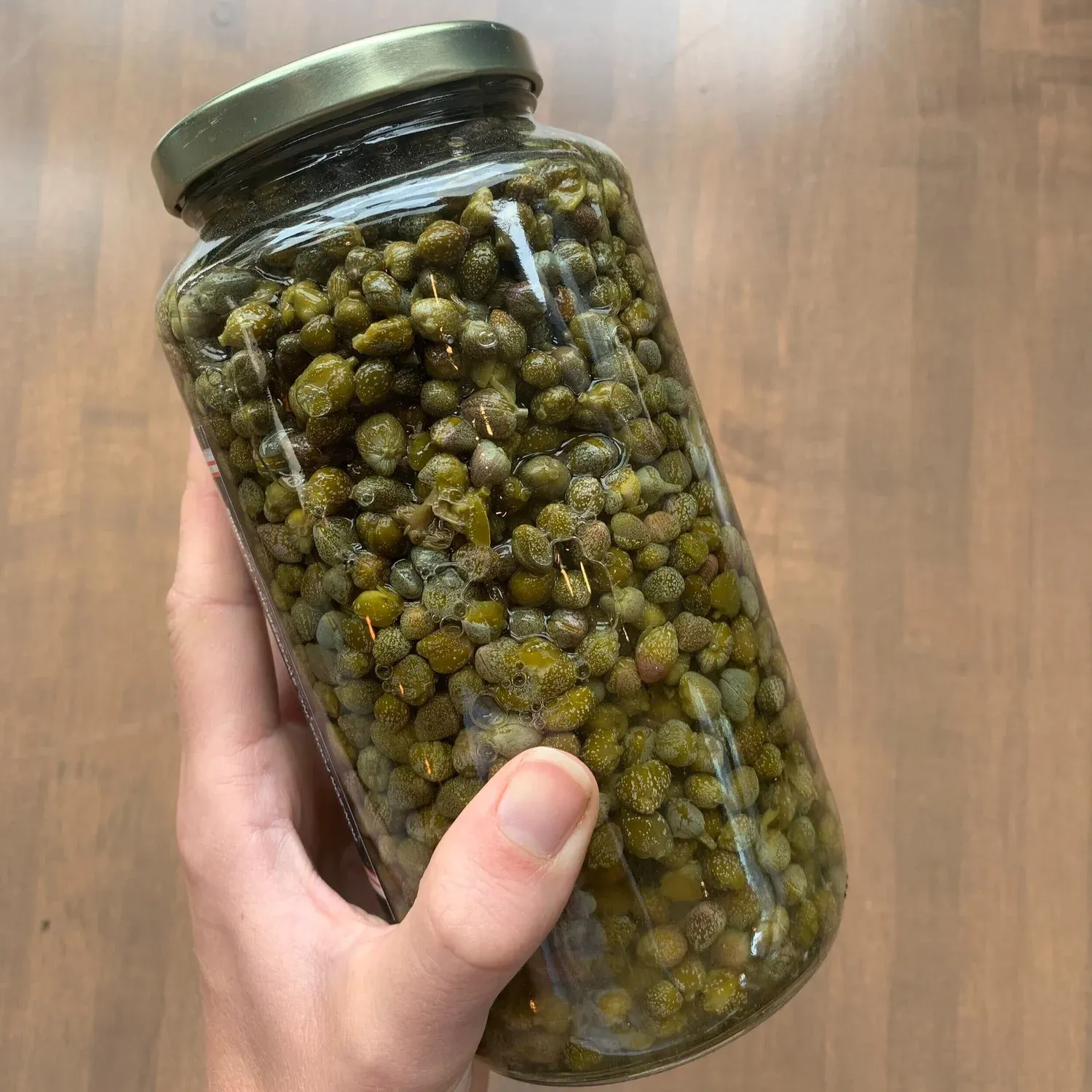 Hand holding a large glass jar packed with brined capers above a wooden table
