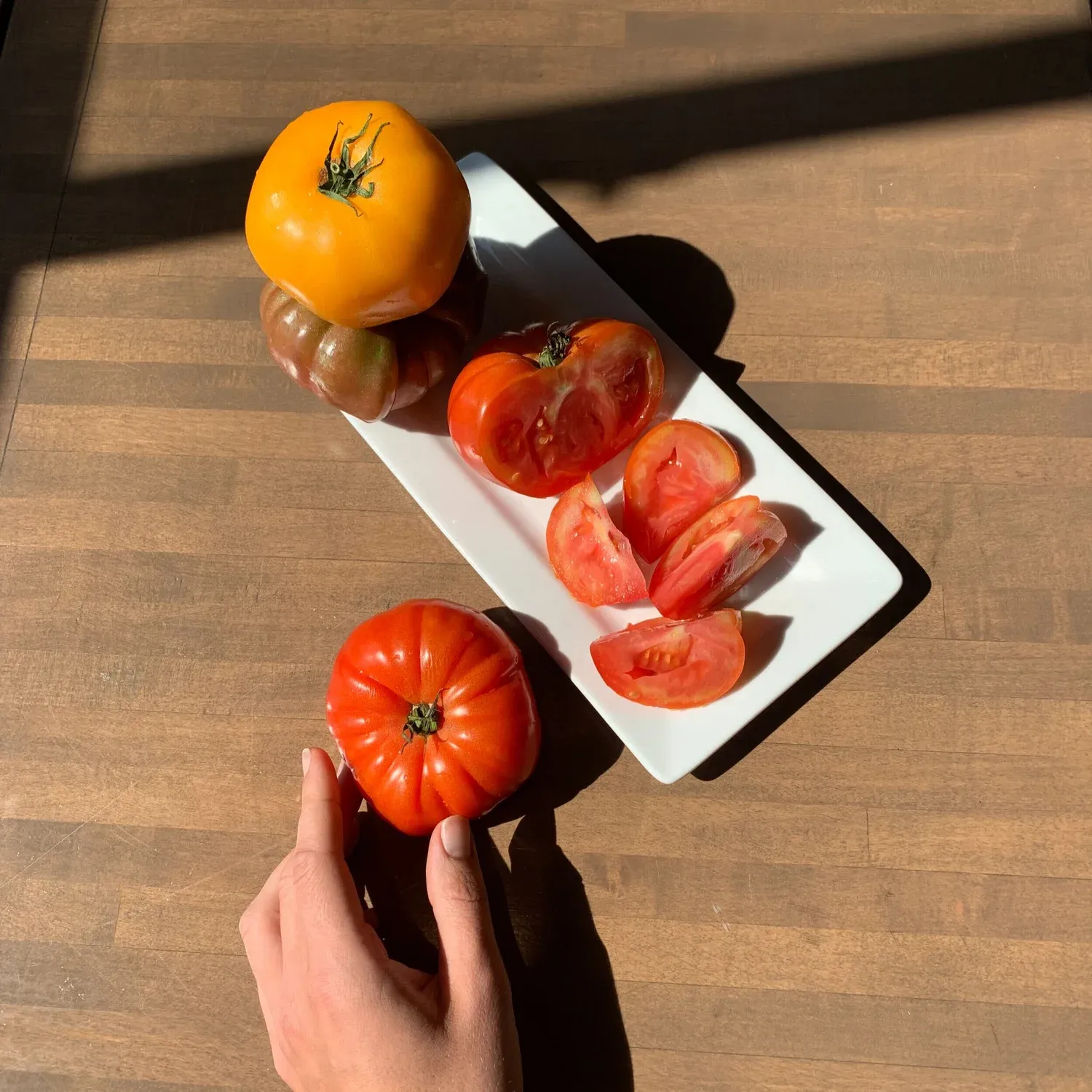 Hand reaching for a red heirloom tomato beside a plate of sliced tomatoes and whole yellow and purple heirlooms