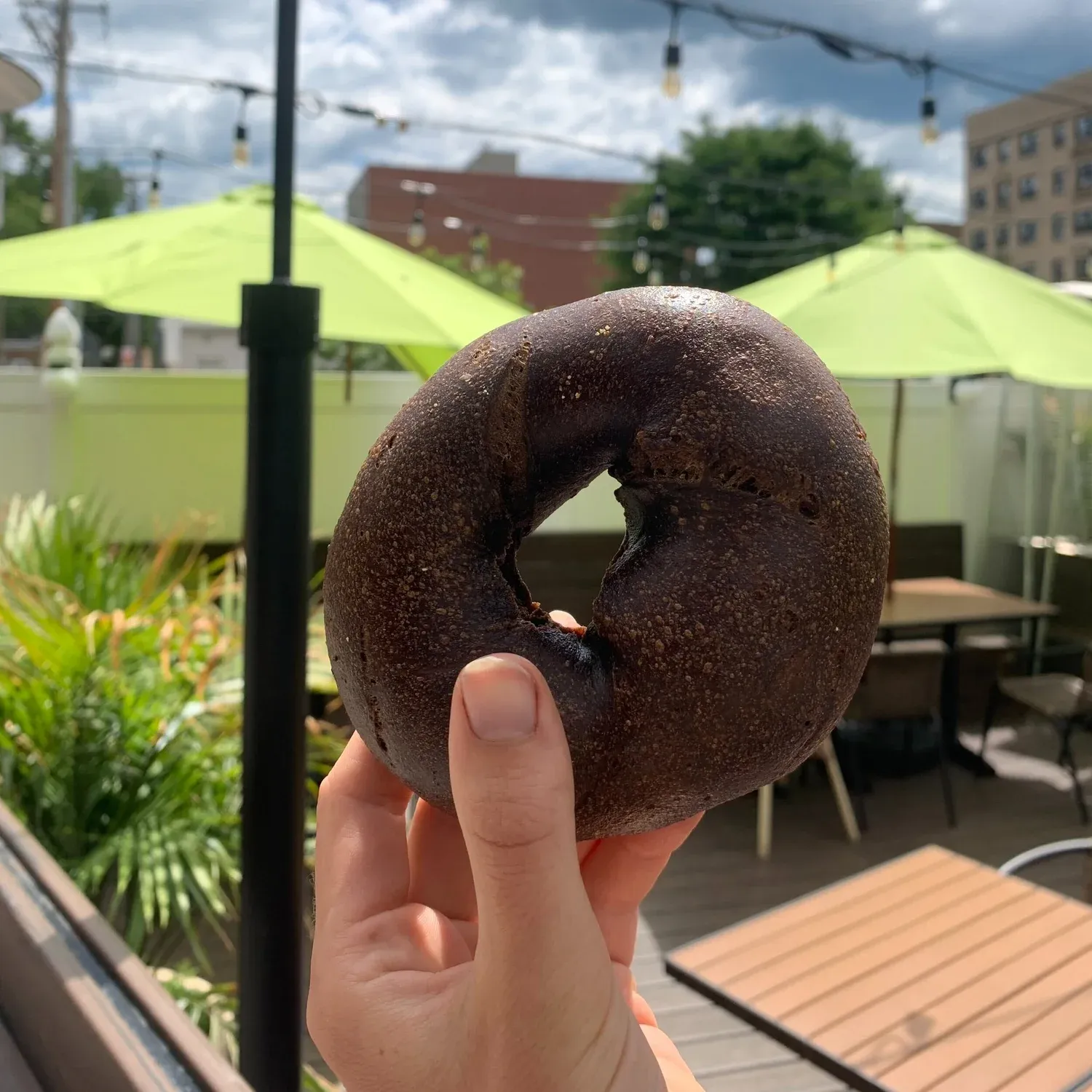 Hand holding a dark pumpernickel bagel on a patio with green umbrellas and string lights
