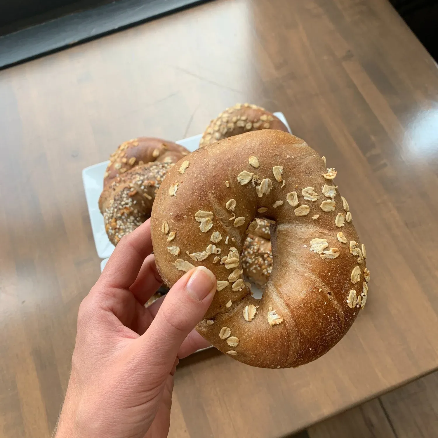 Hand holding an oat-topped whole wheat bagel above a plate of more bagels on a wooden table