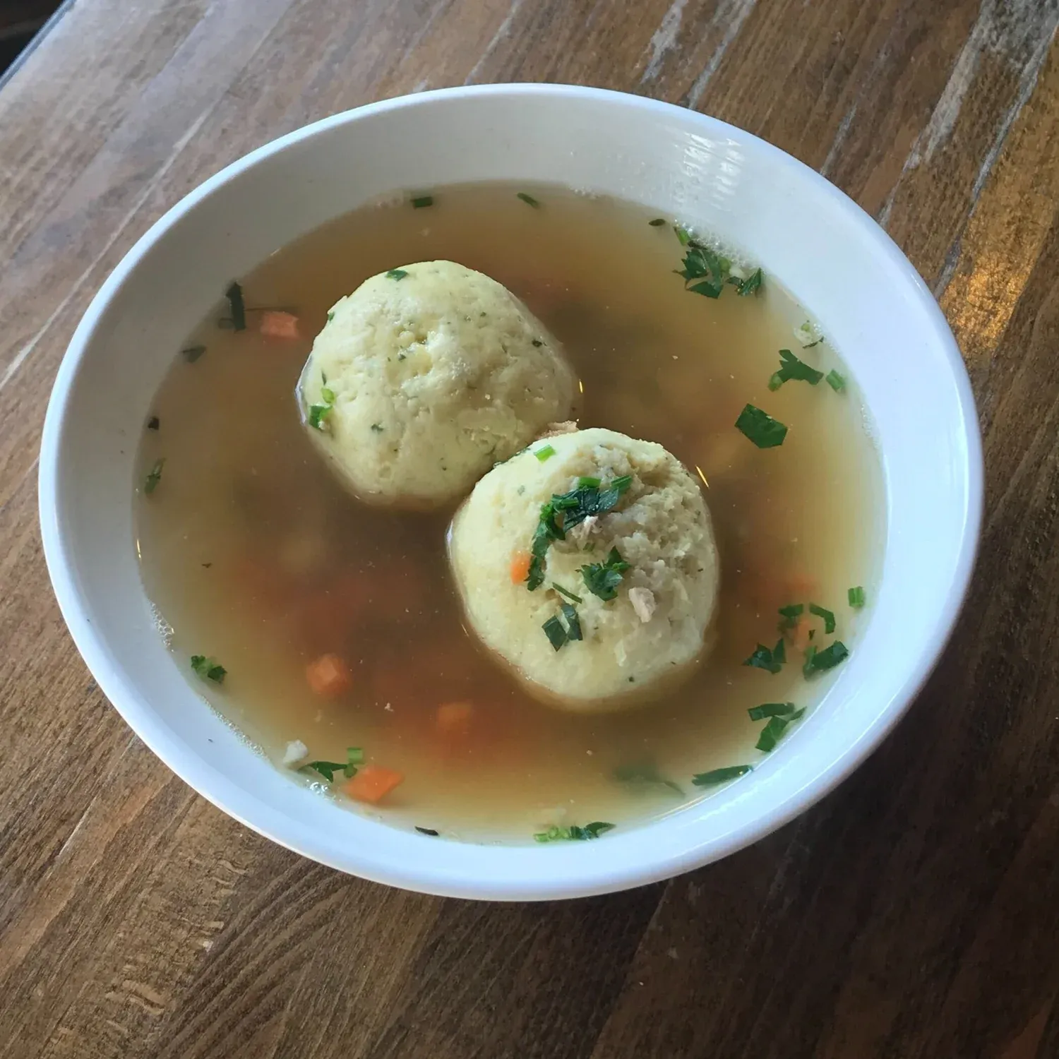 Bowl of clear chicken broth with two matzo balls, diced carrots and chopped parsley